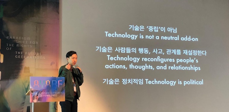 대형 화면 앞에서 마이크를 들고 발표하는 모습. 화면에는 '기술은 중립이 아님 / 기술은 사람들의 행동, 사고, 관계를 재설정한다 / 기술은 정치적임'이라고 적혀 있다.