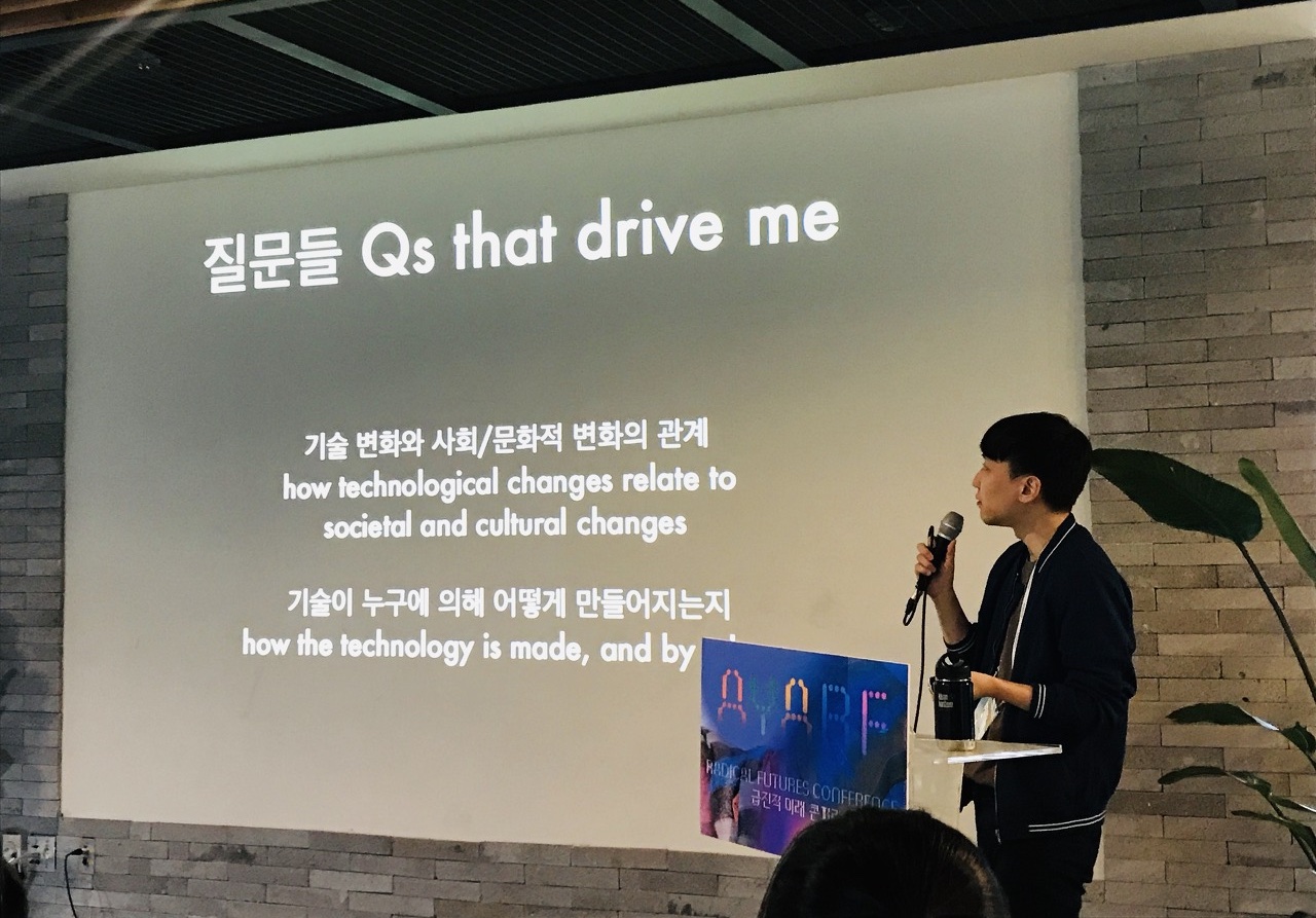 대형 화면 앞에서 마이크를 들고 발표하는 모습. 화면에는 '질문들 / 기술 변화와 사회,문화적 변화의 관계 / 기술이 누구에 의해 어떻게 만들어지는지'라고 적혀 있다.